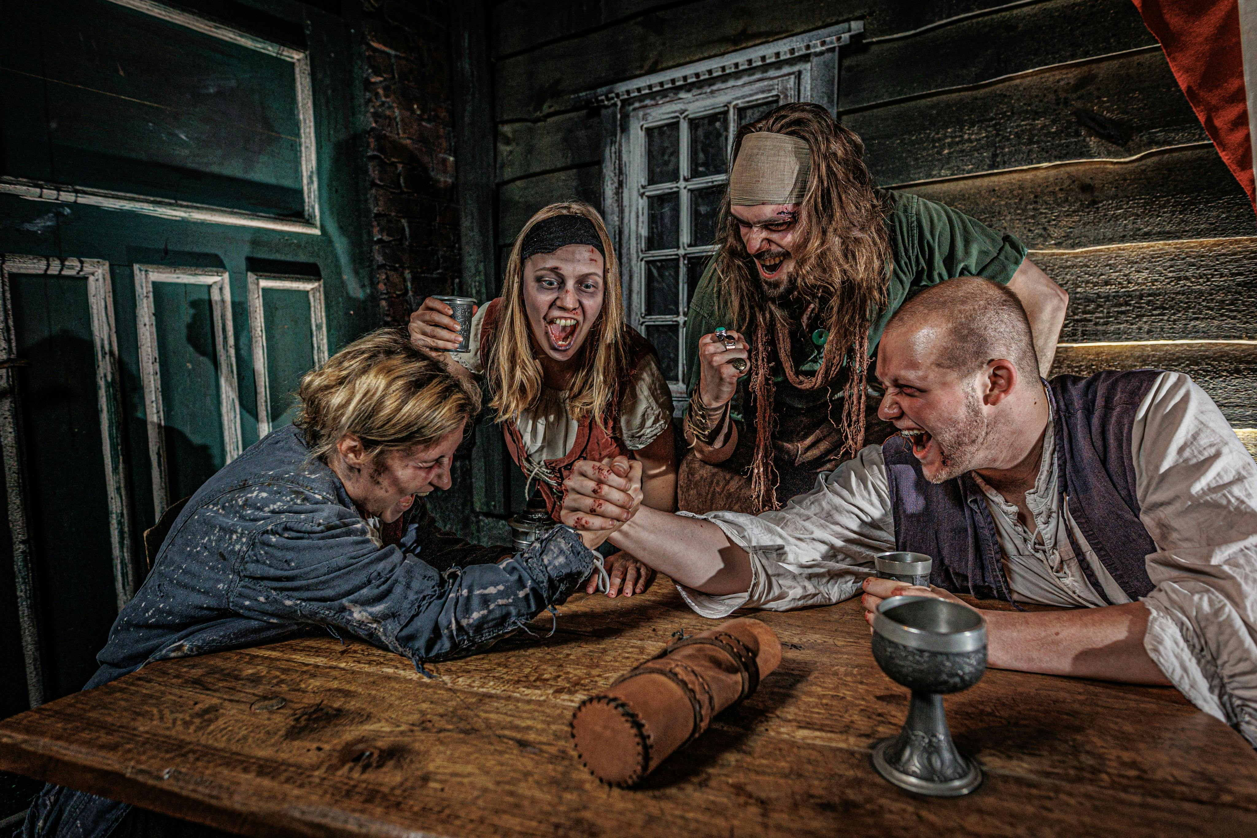 Quatre personnes en tenue rustique se livrant à un bras de fer dans un décor de taverne en bois.