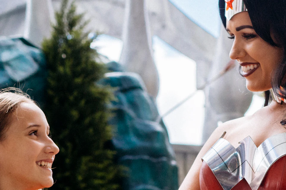 A woman dressed as Wonder Woman smiles at a young girl wearing a matching costume. They stand in front of a themed amusement park ride.