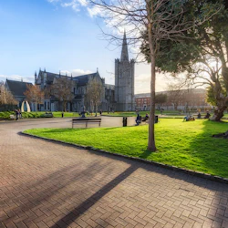 Människor som sitter på bänkar och går i en park med en historisk stenkyrka och frodig grönska under en delvis molnig himmel.