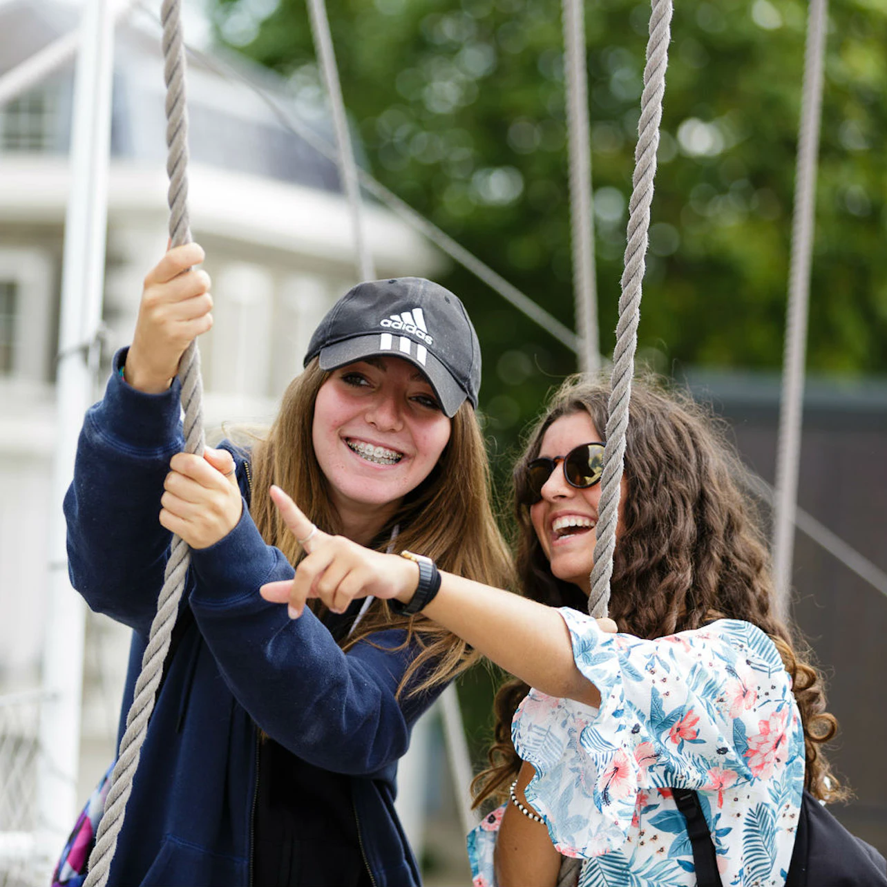 Two smiling young women, one in a cap with braces and the other in sunglasses, holding ropes outdoors, in a casual setting.