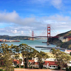 View of the Golden Gate Bridge with water, trees, and buildings in the foreground under a blue sky with clouds.
