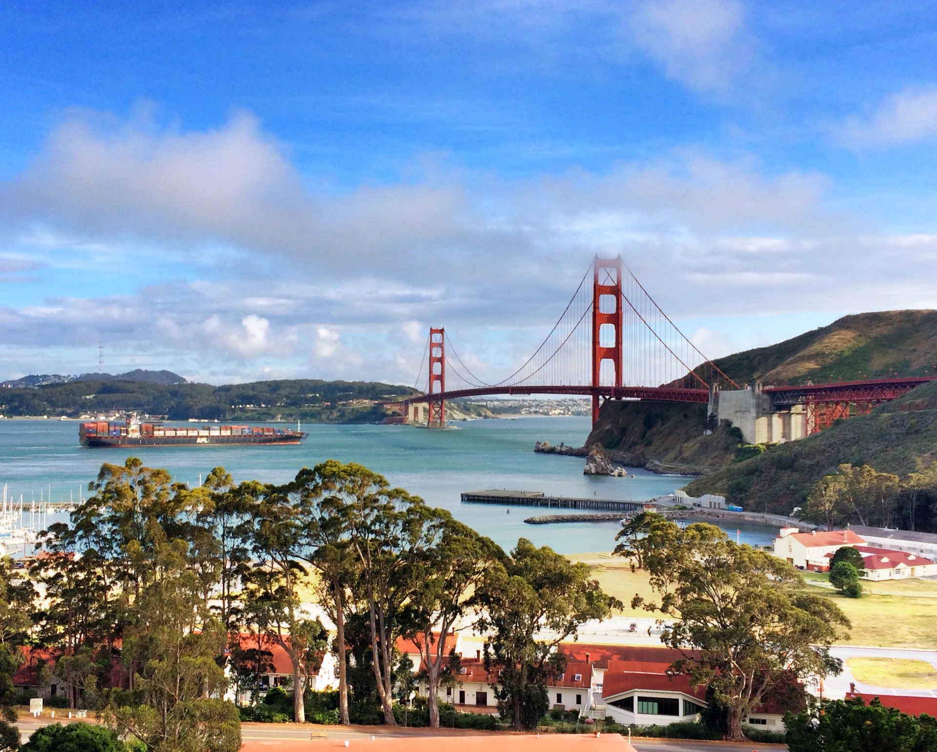 View of the Golden Gate Bridge with water, trees, and buildings in the foreground under a blue sky with clouds.