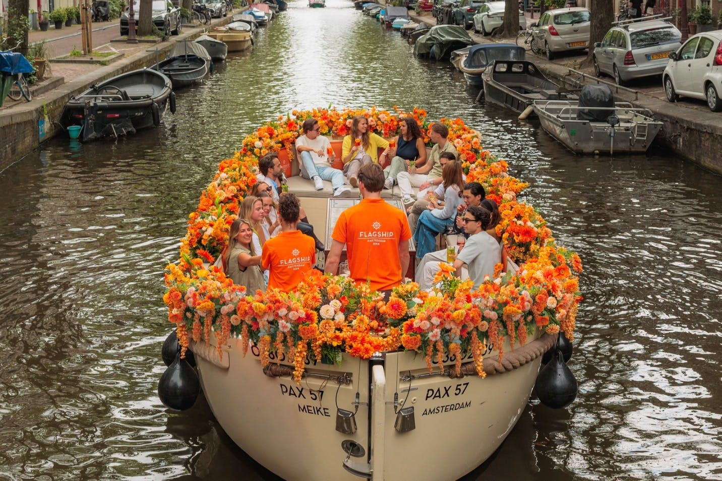 A boat decorated with orange flowers carrying people on a canal, surrounded by parked cars and bicycles.