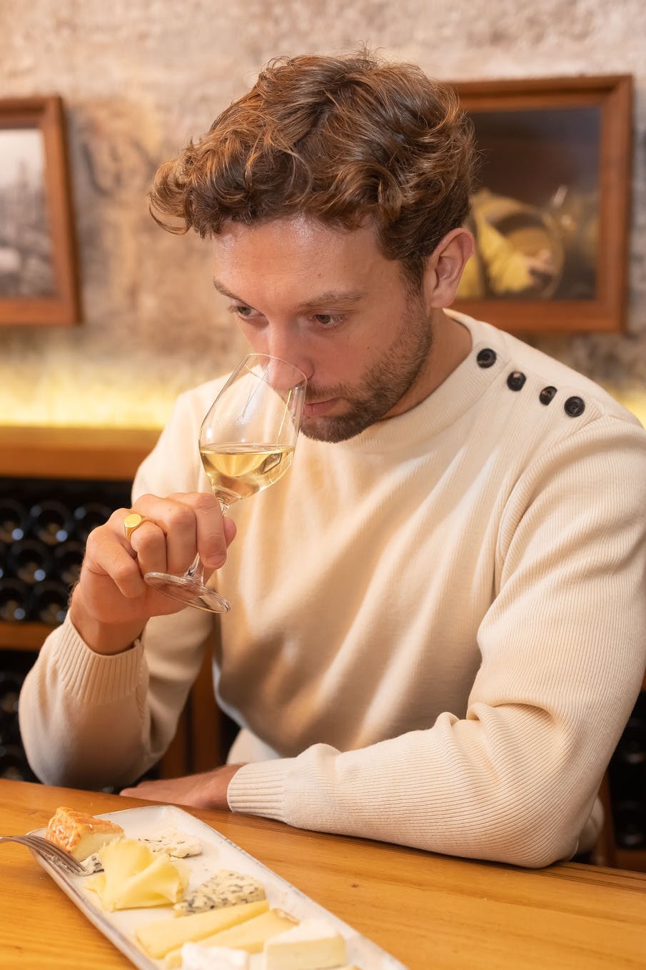 베이지색 스웨터를 입은 남성이 벽에 액자 그림이 그려진 아늑한 분위기에서 화이트 와인 한 잔의 냄새를 맡고 있습니다.