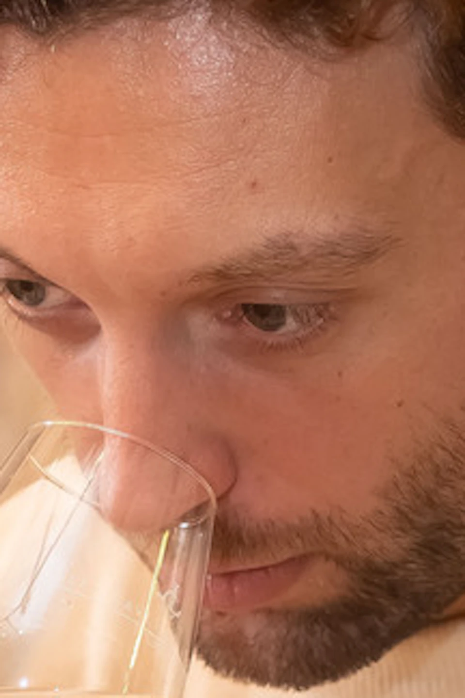 A man in a beige sweater smelling a glass of white wine in a cozy setting with framed pictures on the wall.