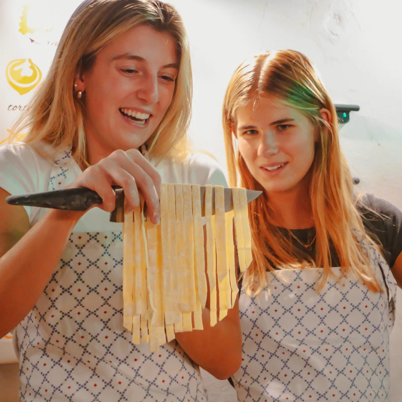 Two women wearing aprons are smiling and holding freshly cut pasta strips in a cooking class setting.