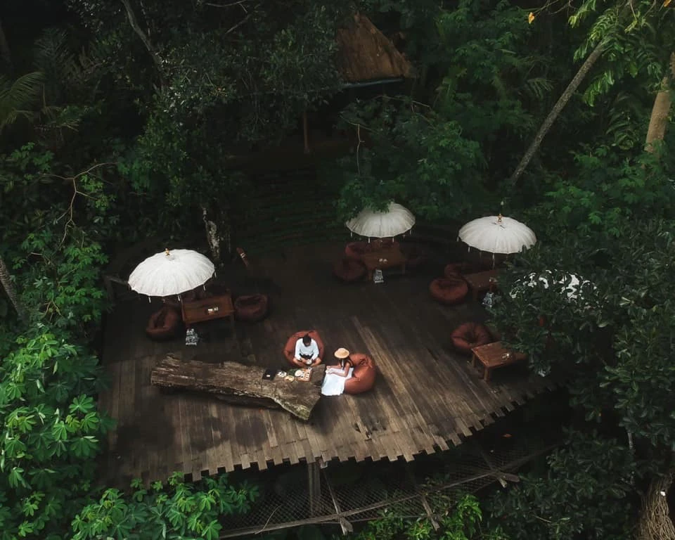 Aerial view of a wooden deck in a forest with tables, chairs, white parasols, and two people sitting by a large wooden table.