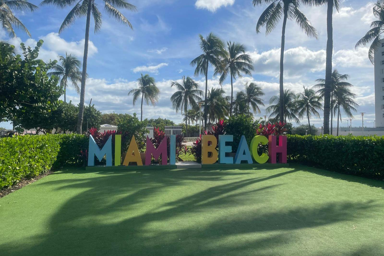 Large, colorful letters spelling "MIAMI BEACH" on a grassy area with palm trees and a blue sky in the background.