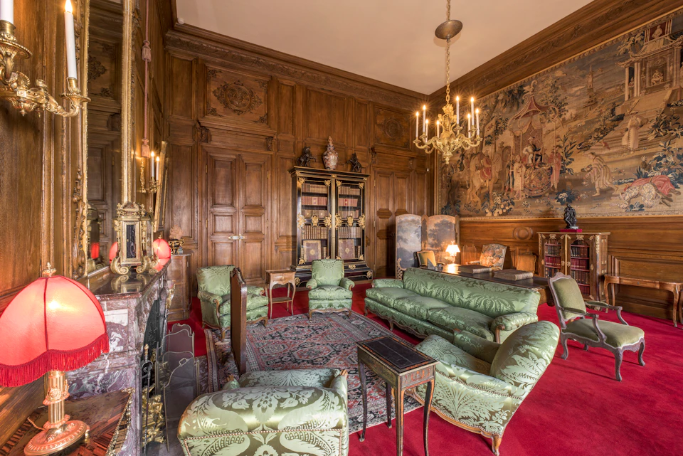 A luxurious room with wood-paneled walls, green upholstered furniture, a large bookcase, ornate chandeliers, and a detailed tapestry.
