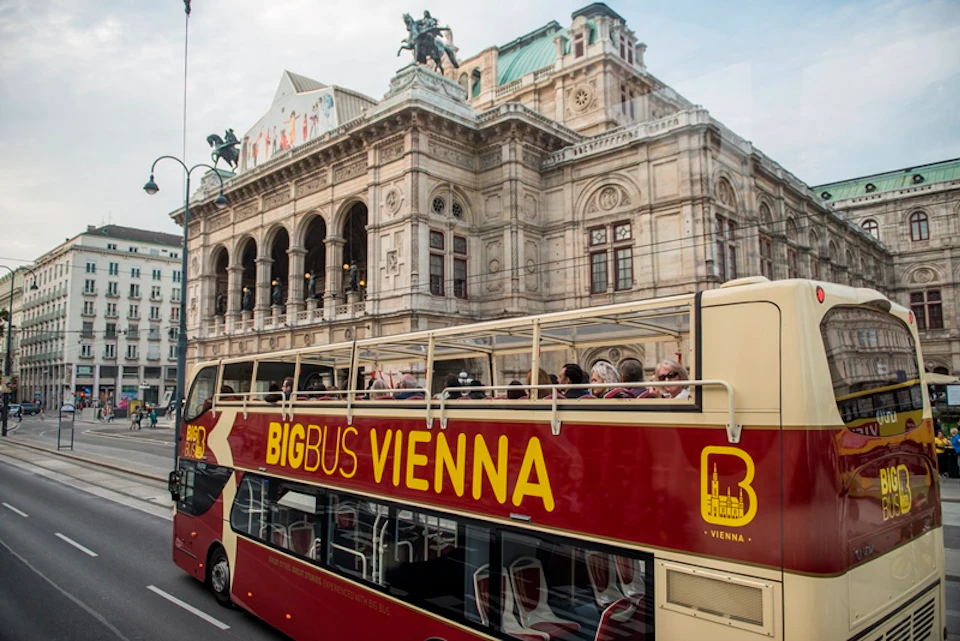 A double-decker bus labeled "Big Bus Vienna" passes by a historic building with arched windows and sculptures on its roof.