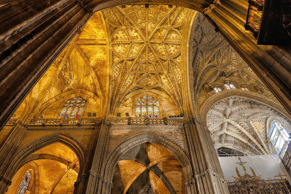 Intricate vaulted ceiling of a gothic cathedral with ornate details and stained glass windows.
