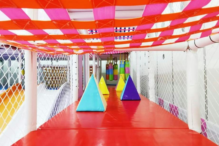 A colorful indoor play area with pyramid-shaped obstacles on a red floor, netted walls, and a checkered ceiling pattern.