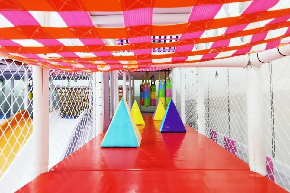 A colorful indoor play area with pyramid-shaped obstacles on a red floor, netted walls, and a checkered ceiling pattern.