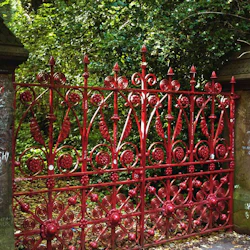 Röd utsmyckad grind med klotterklädda pelare på båda sidor, omgiven av täta gröna blad. ”Strawberry Field” står skrivet på vänster pelare.