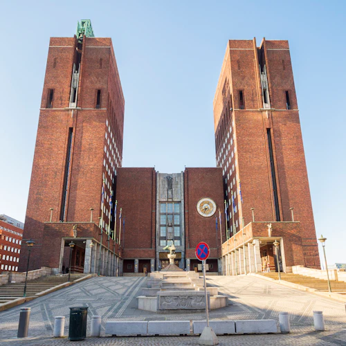 En stor tegelbyggnad med två höga torn, breda trappsteg och en parkeringsförbudsskylt framför. Flaggor och ett cirkulärt emblem är synliga.