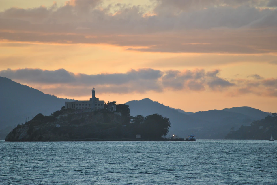 An island with a large building and lighthouse, silhouetted against a sunset sky, surrounded by calm water and distant hills.