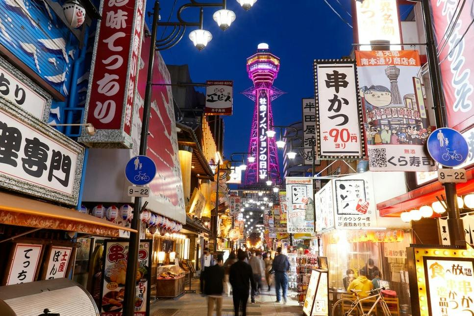 A busy pedestrian street at night with colorful neon signs and a lit-up tower in the background.