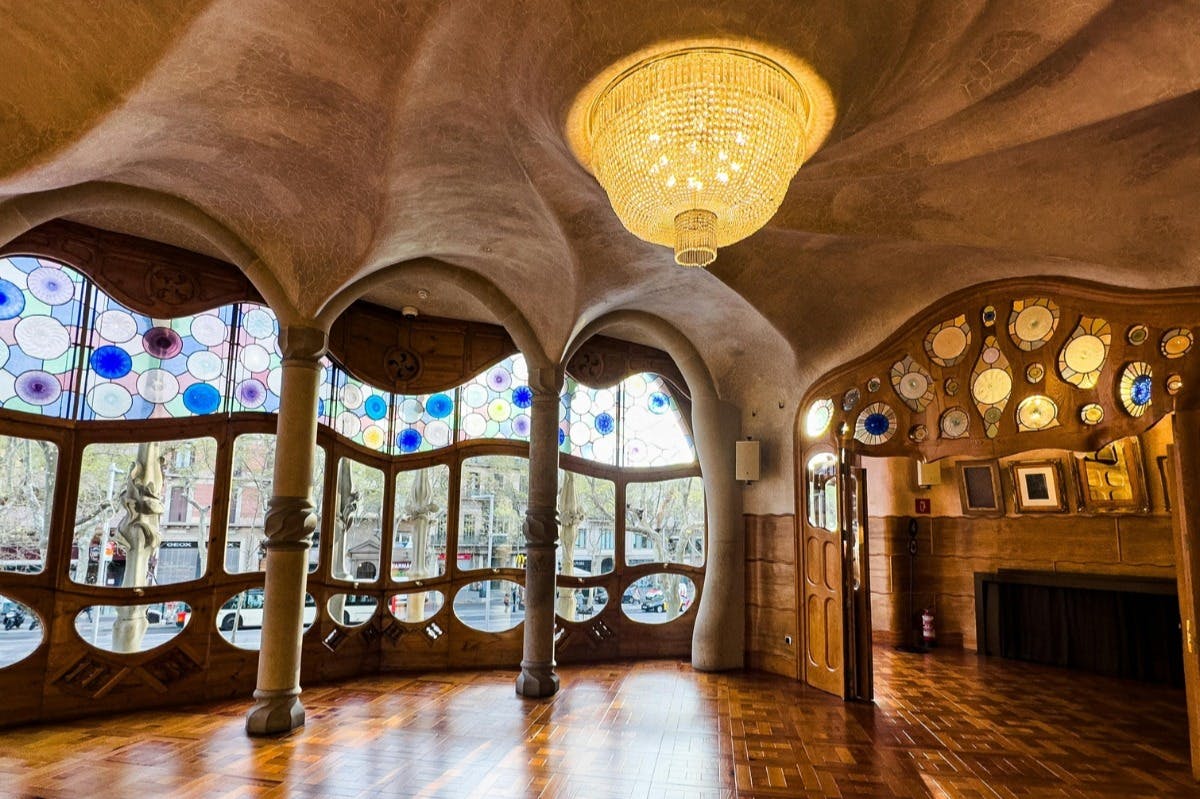 Sierlijke kamer met boogramen, gekleurd glas, houten vloer en een grote kroonluchter die aan een gebeeldhouwd plafond hangt.