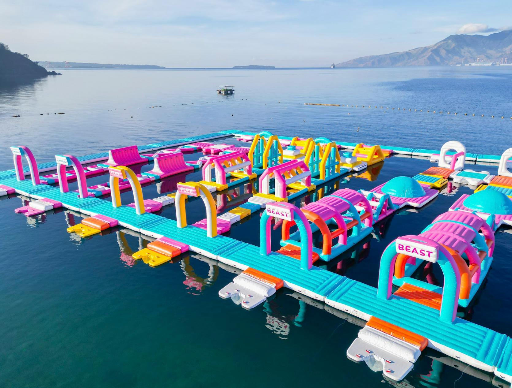 A colorful floating inflatable obstacle course on a calm lake with a scenic mountain backdrop under a clear sky.