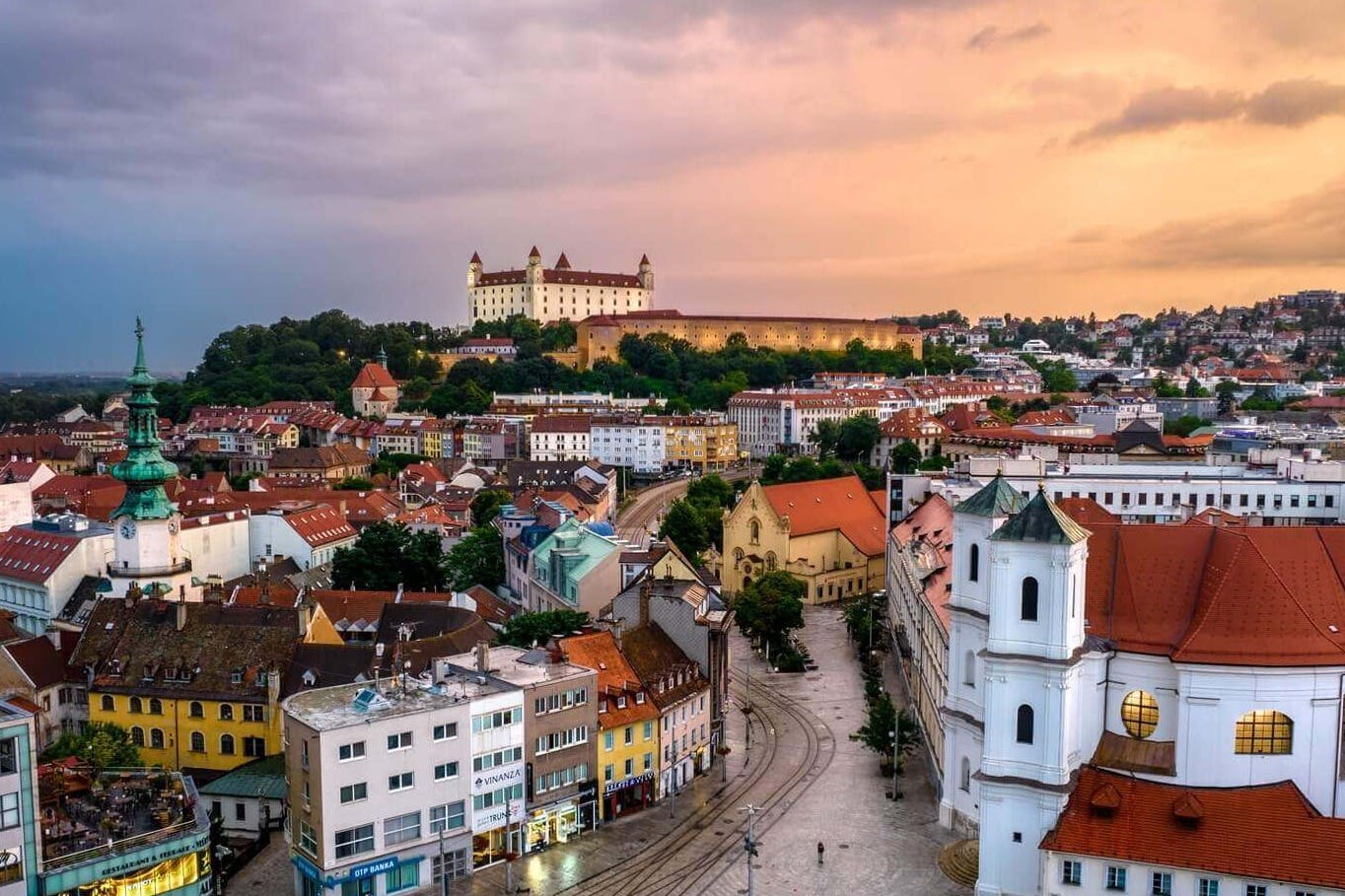 Ένα αστικό τοπίο στο ηλιοβασίλεμα με ένα κάστρο σε έναν λόφο στο βάθος, που περιβάλλεται από πολύχρωμα κτίρια, δρόμους και μια λευκή εκκλησία.