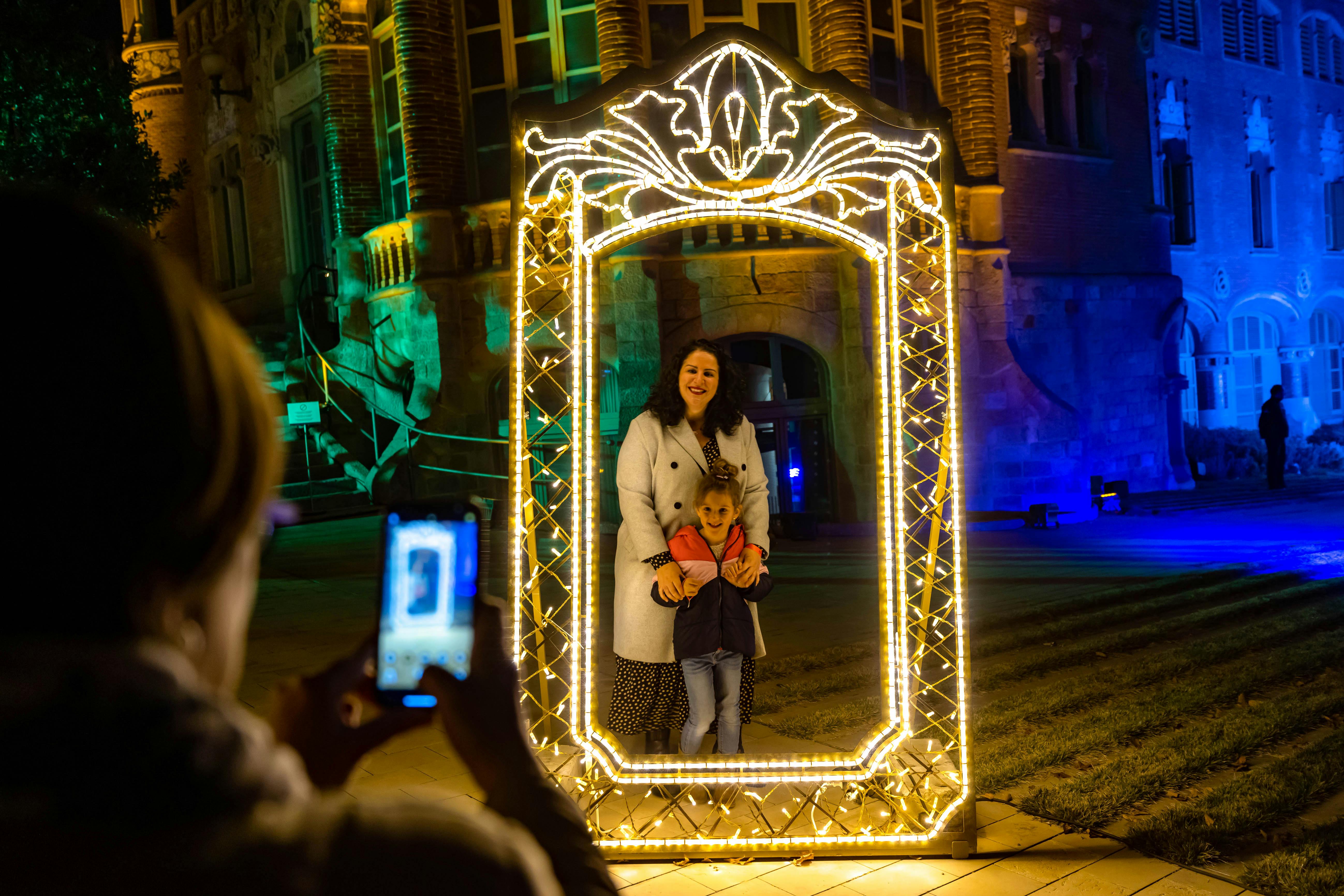 Une femme et un enfant posent à l'intérieur d'un cadre décoratif illuminé alors qu'ils sont photographiés sur un site extérieur éclairé la nuit.