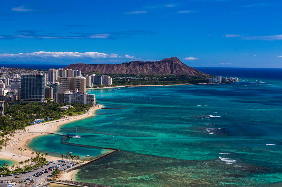 View of a coastline with tall buildings, clear blue water, several boats, and a mountainous backdrop under a blue sky.