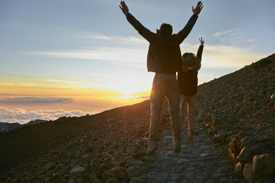 Two people with raised arms hike on a rocky path at sunset, with a scenic view of the sky and distant clouds.