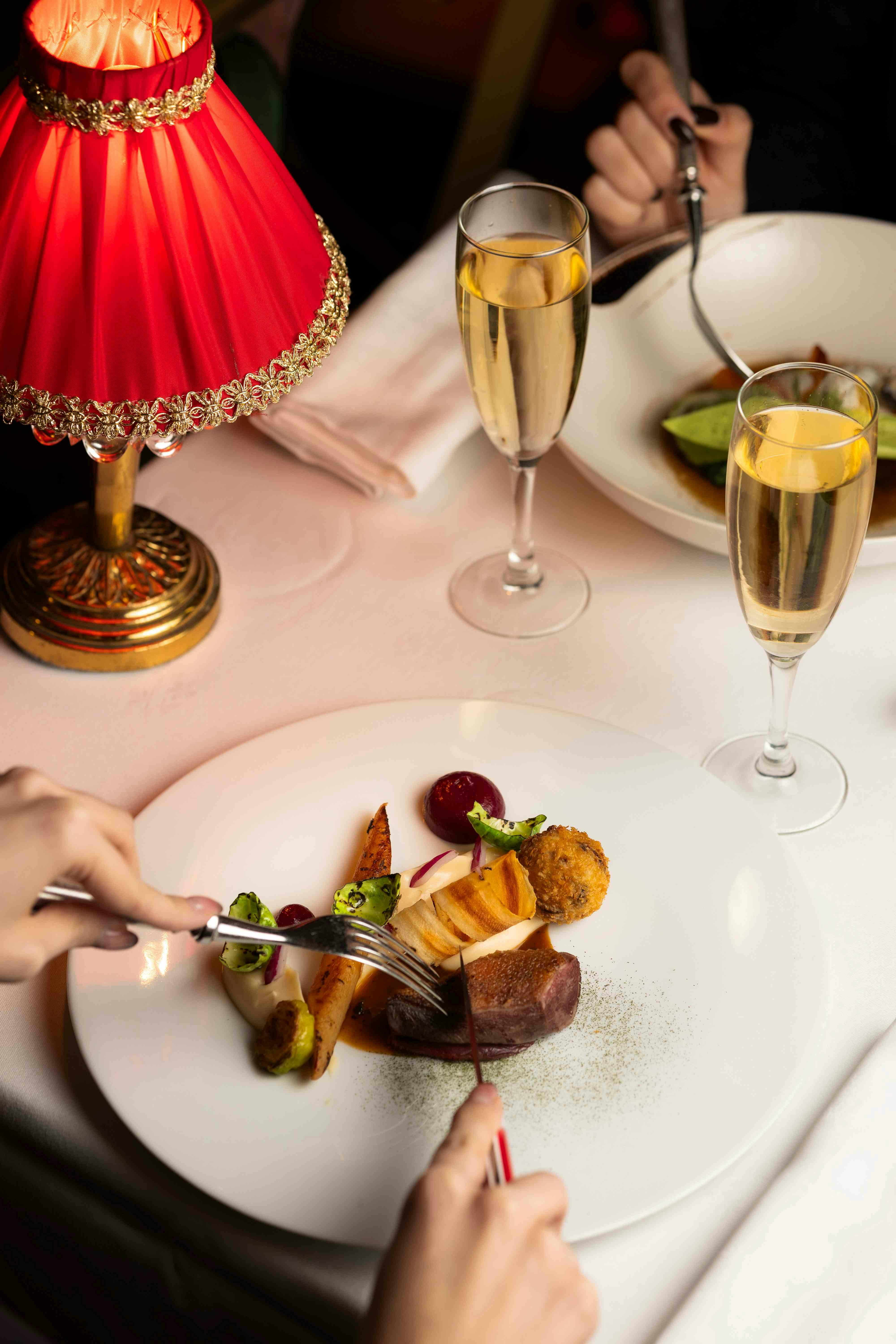 A table setting with a red lamp, two glasses of champagne, and plates of food, with hands holding utensils visible.
