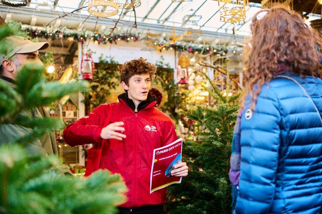 A man in a red jacket holds a brochure and talks to another man among Christmas decorations and trees in a festive indoor setting.