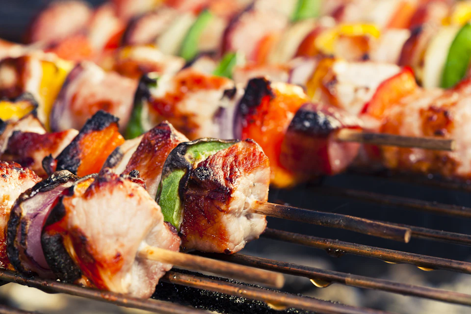 Close-up of barbequed skewers with grilled meat, red and green bell peppers, and yellow squash on a grill.