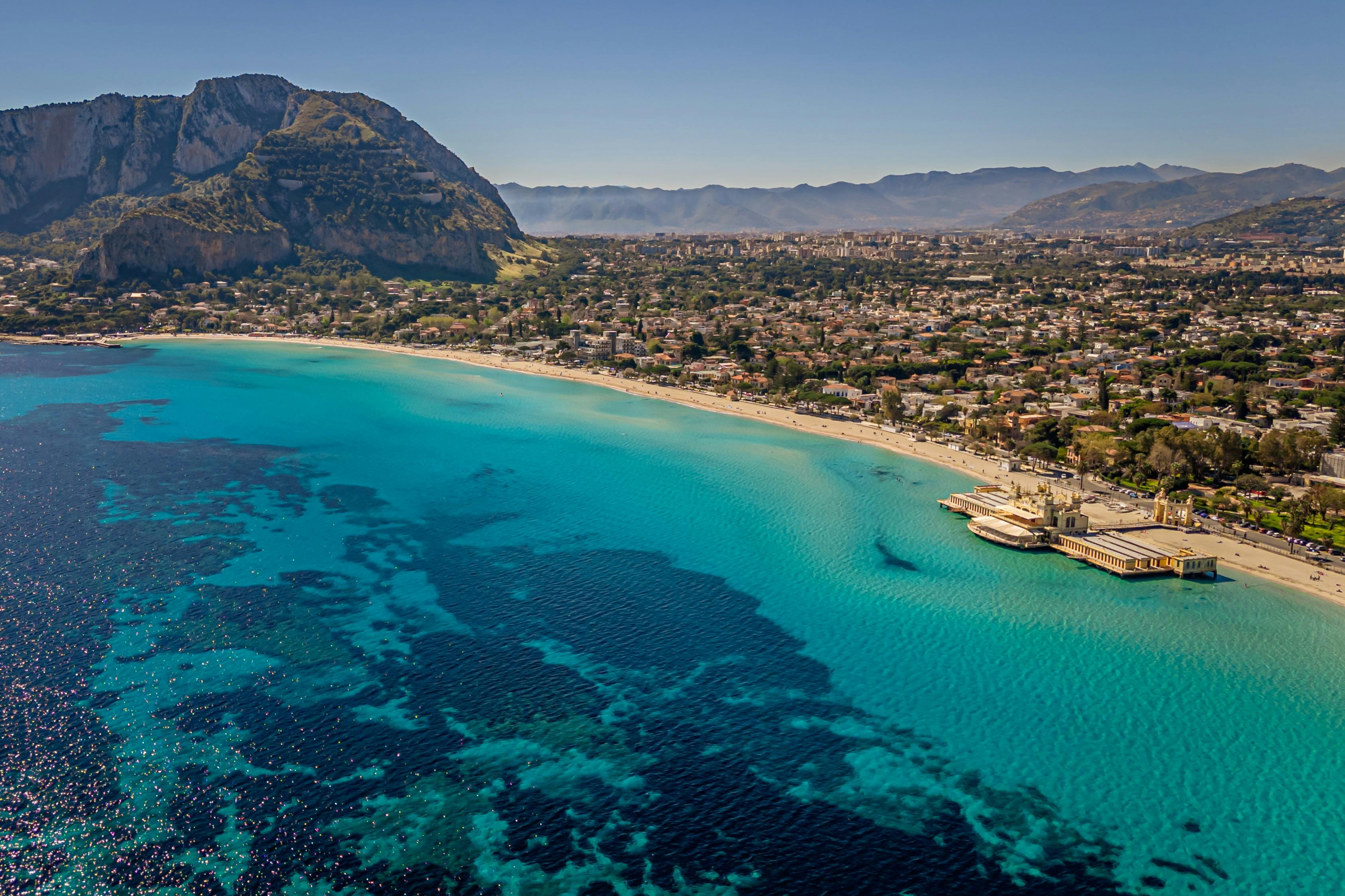 Vista aerea di una città costiera con acqua turchese, spiaggia sabbiosa, numerosi edifici e montagne sullo sfondo.