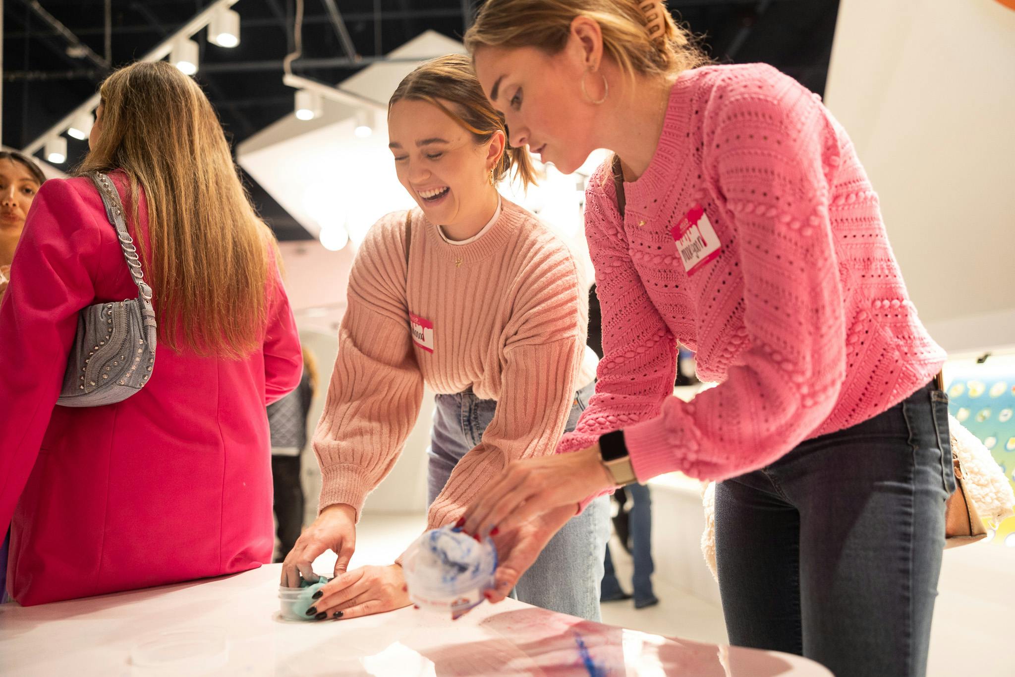 To kvinder i lyserøde trøjer deltager i en kunstaktivitet på et bord, mens de smiler. En menneskemængde er synlig i baggrunden.