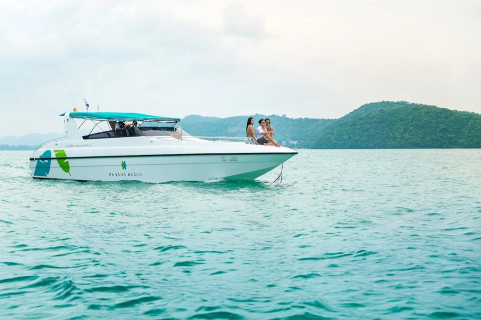 Three people sitting at the bow of a white boat named "Banana Beach" in calm turquoise water with green hills in the background.