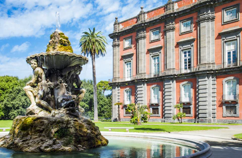 A historic red building with large windows next to a decorative fountain surrounded by greenery and a palm tree.