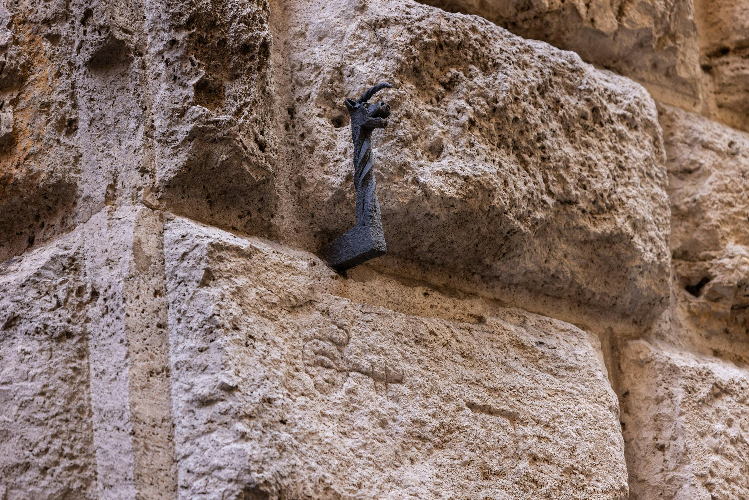 Una piccola figura metallica contorta che assomiglia a un drago è montata su una parete di pietra grezza.