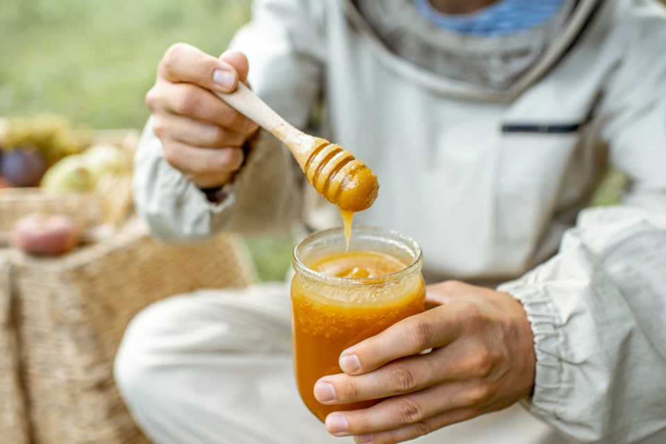 A person in a beekeeping suit holding a jar of honey and dipping a honey dipper into it outdoors.