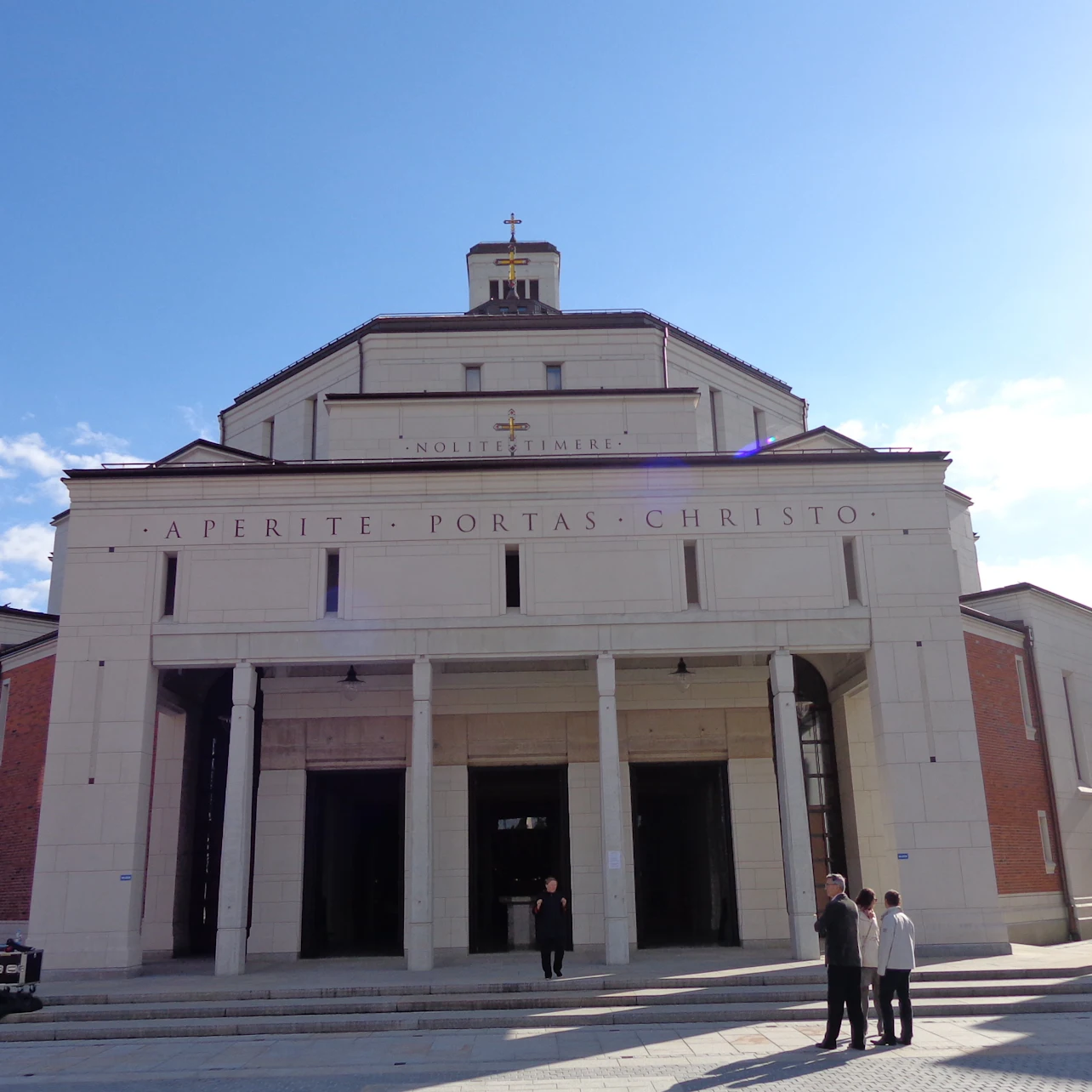 A church facade with columns and a cross on top. Four people are standing in front under a clear blue sky.