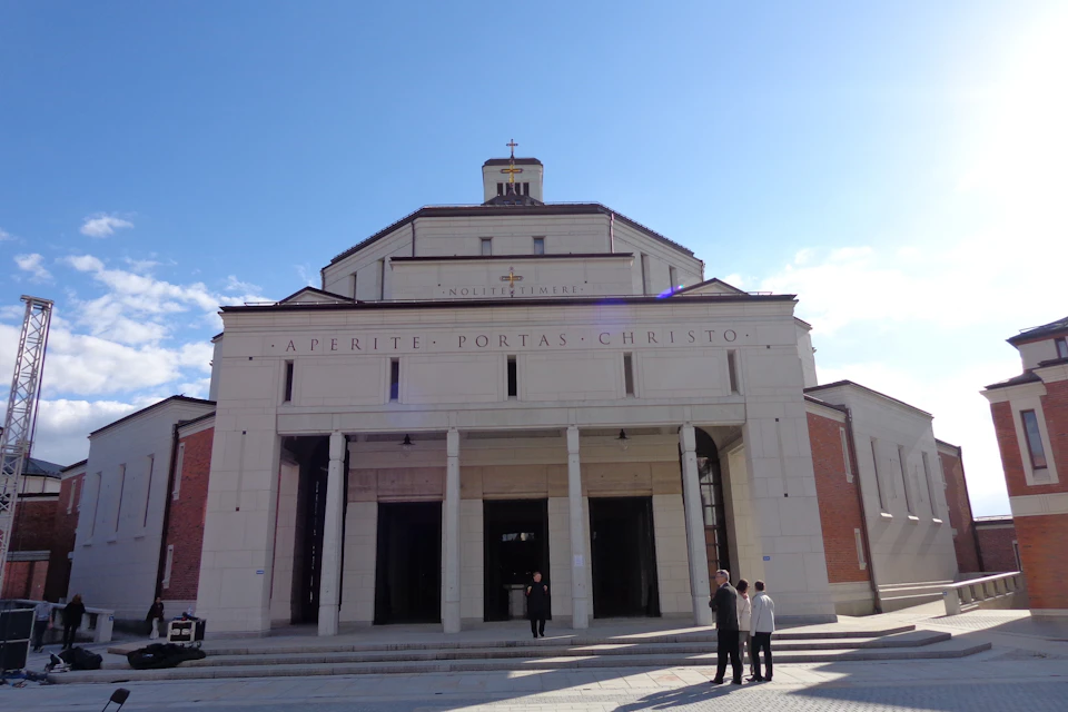 A church facade with columns and a cross on top. Four people are standing in front under a clear blue sky.
