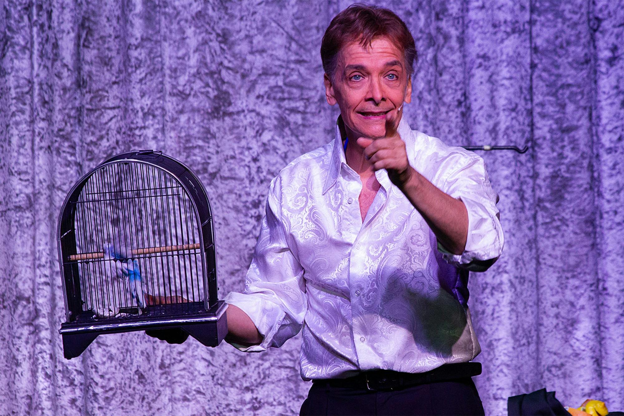 A performer in a white shirt gestures onstage, holding a birdcage, with a textured purple curtain as the background.