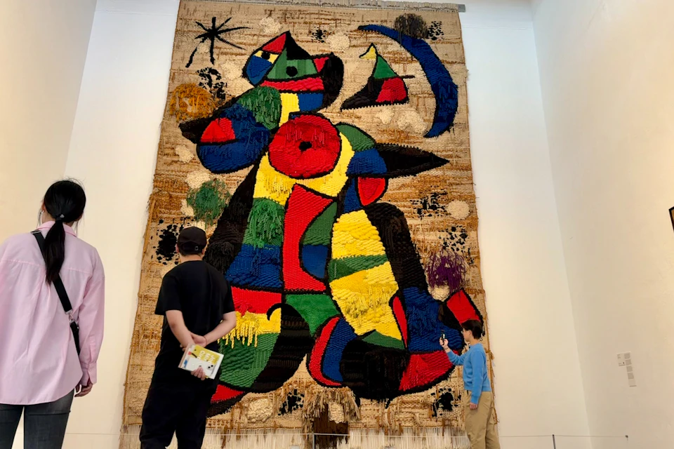 Two people observe a large, colorful abstract tapestry mounted on a gallery wall, featuring bold shapes and textures.