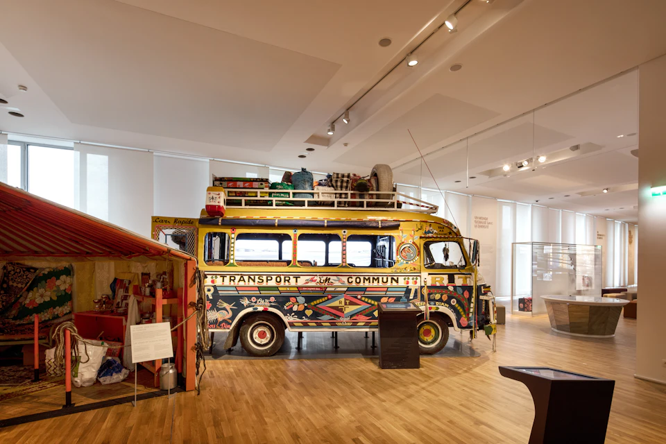 A decorated vintage bus with luggage on top, displayed in a brightly lit museum room with wooden floors.