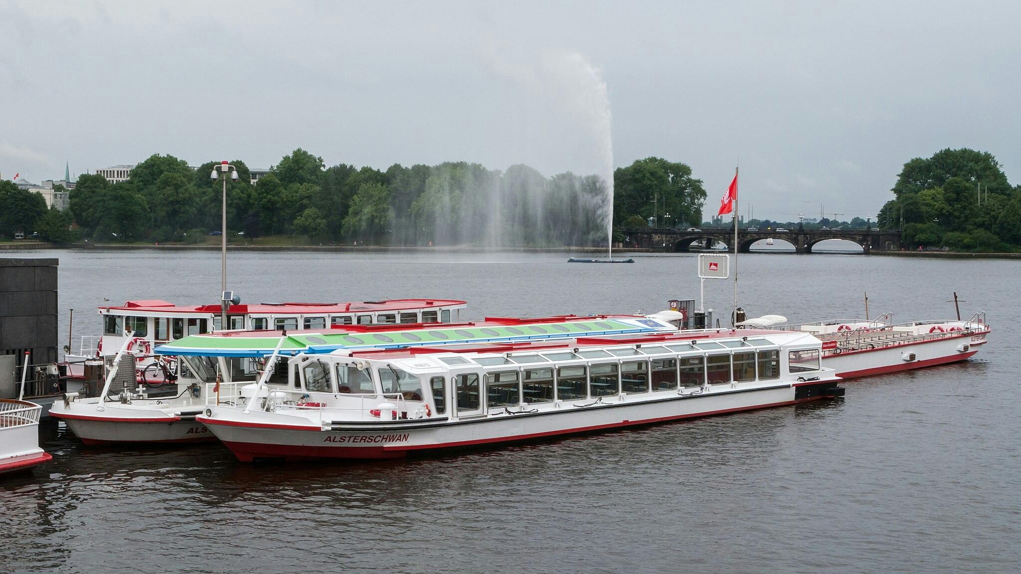 Vita och röda båtar dockade på en flod med en fontän i fjärran, omgiven av grönskande träd och en bro synlig i bakgrunden.