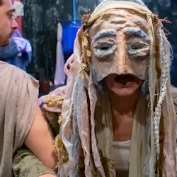 En person i teaterkostym och mask med tygremsor, flankerad av två andra i liknande klädsel på en mörk scen.