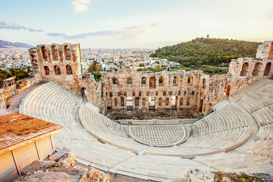 Ancient stone amphitheater with tiered seating, overlooking a modern city and distant hills under a partly cloudy sky.