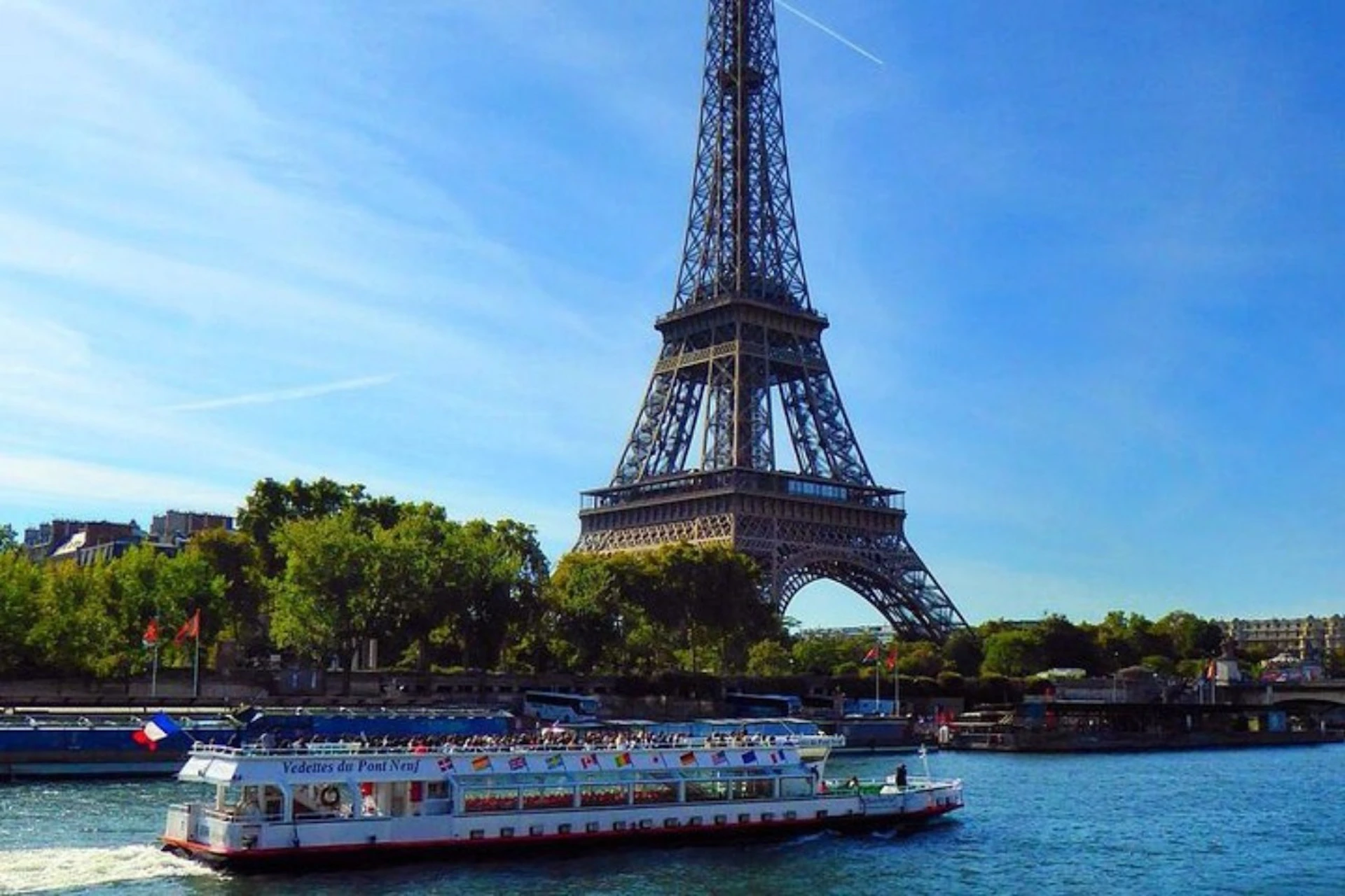 A sightseeing boat on a river in front of the Eiffel Tower on a sunny day with trees and blue sky in the background.