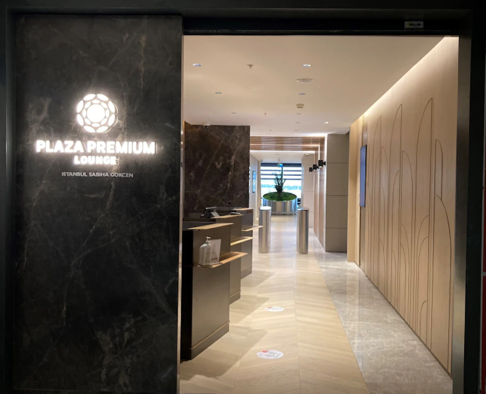 Entrance of Plaza Premium Lounge with modern decor, illuminated sign, marble walls, and a view of seating area and plants through a hallway.