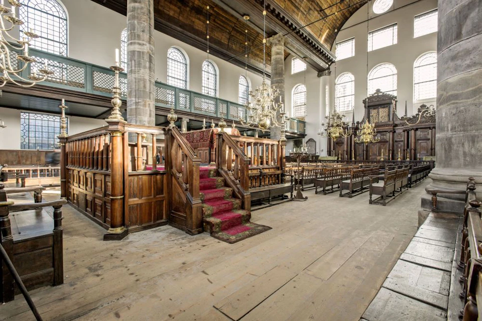 A historic church interior with wooden pews, ornate chandeliers, a red-carpeted pulpit, and large arched windows.