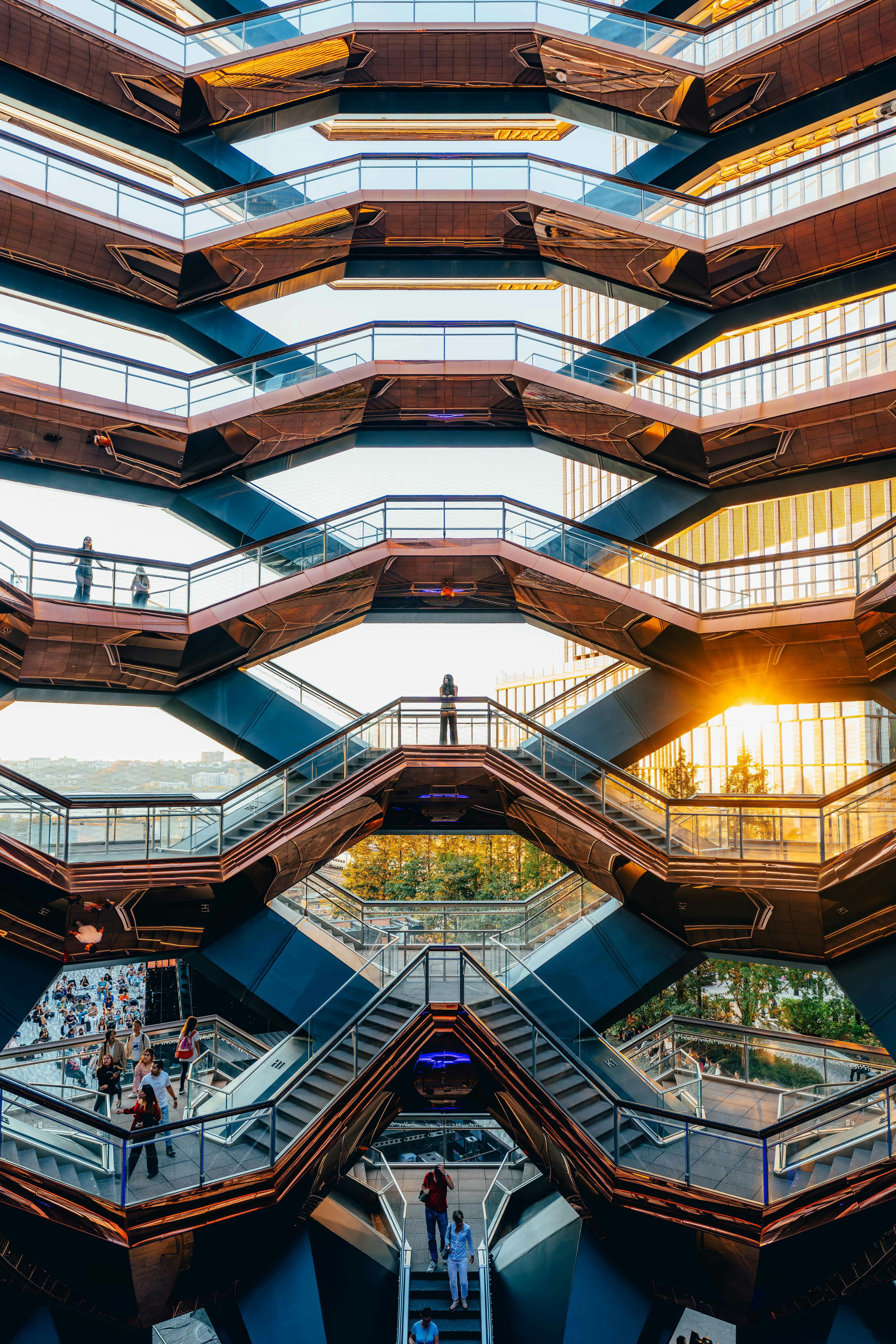 Bronze-colored staircase structure with multiple levels and people walking, against the backdrop of buildings and sunset.