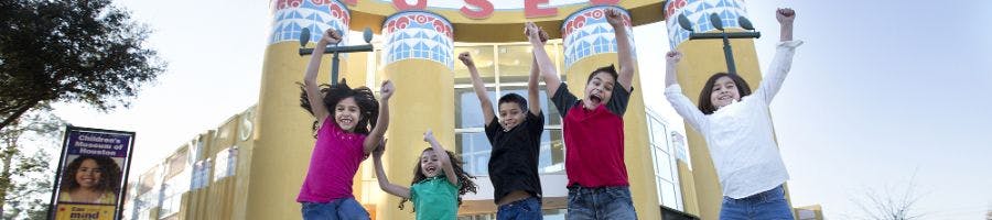 Пятеро детей радостно прыгают с поднятыми руками перед разноцветным зданием. На боковой стороне висит табличка с фотографией девушки.