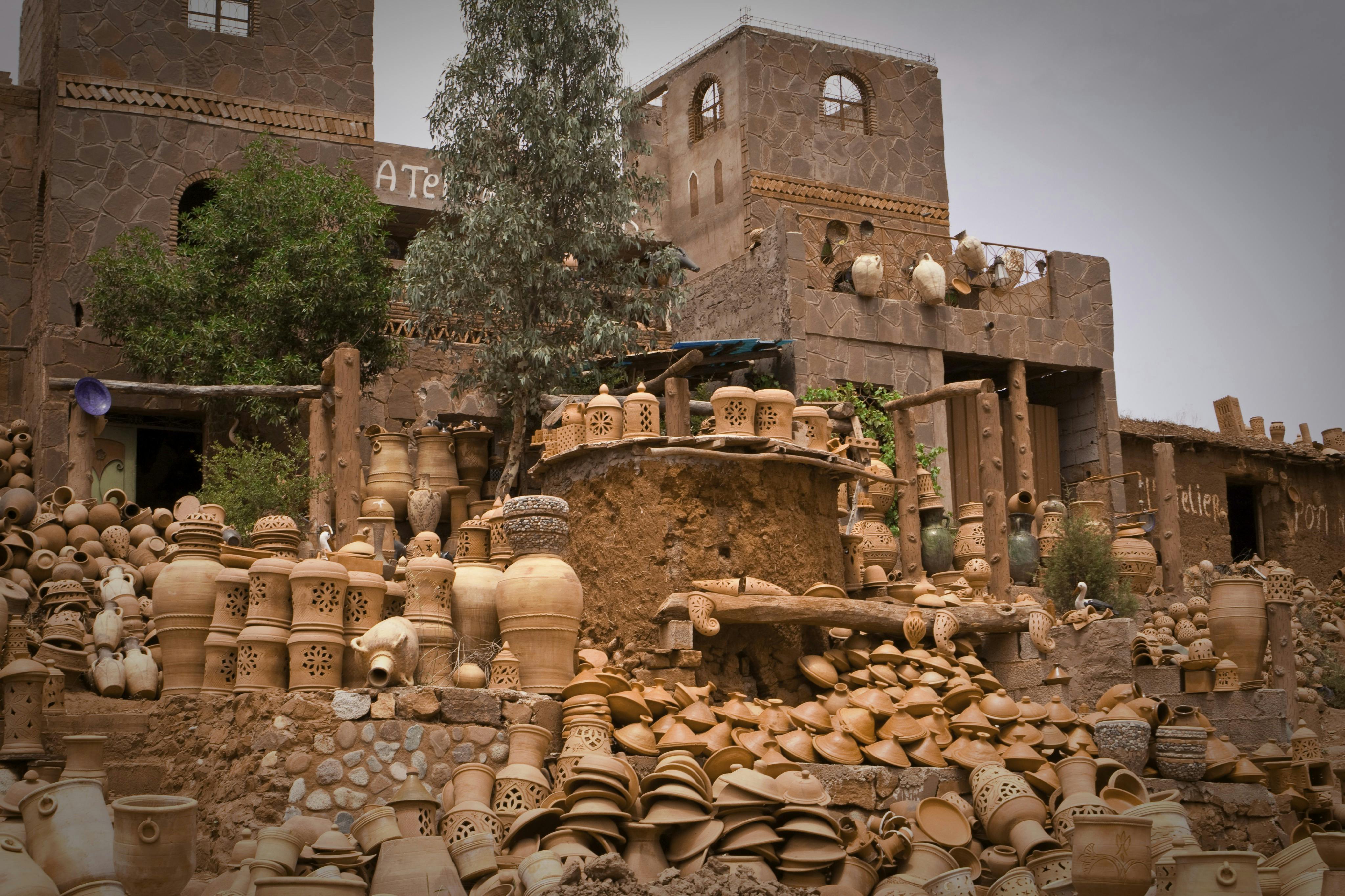 Outdoor pottery workshop featuring numerous clay pots, jugs, and lanterns, set against a rustic stone building with trees nearby.